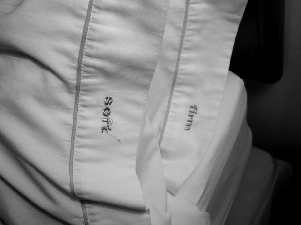 black and white photograph showing a closeup of two hotel pillows, one on top of the other. The pillowcases have embroidered words on the edges. The top pillow says "soft" and the bottom one says "firm"